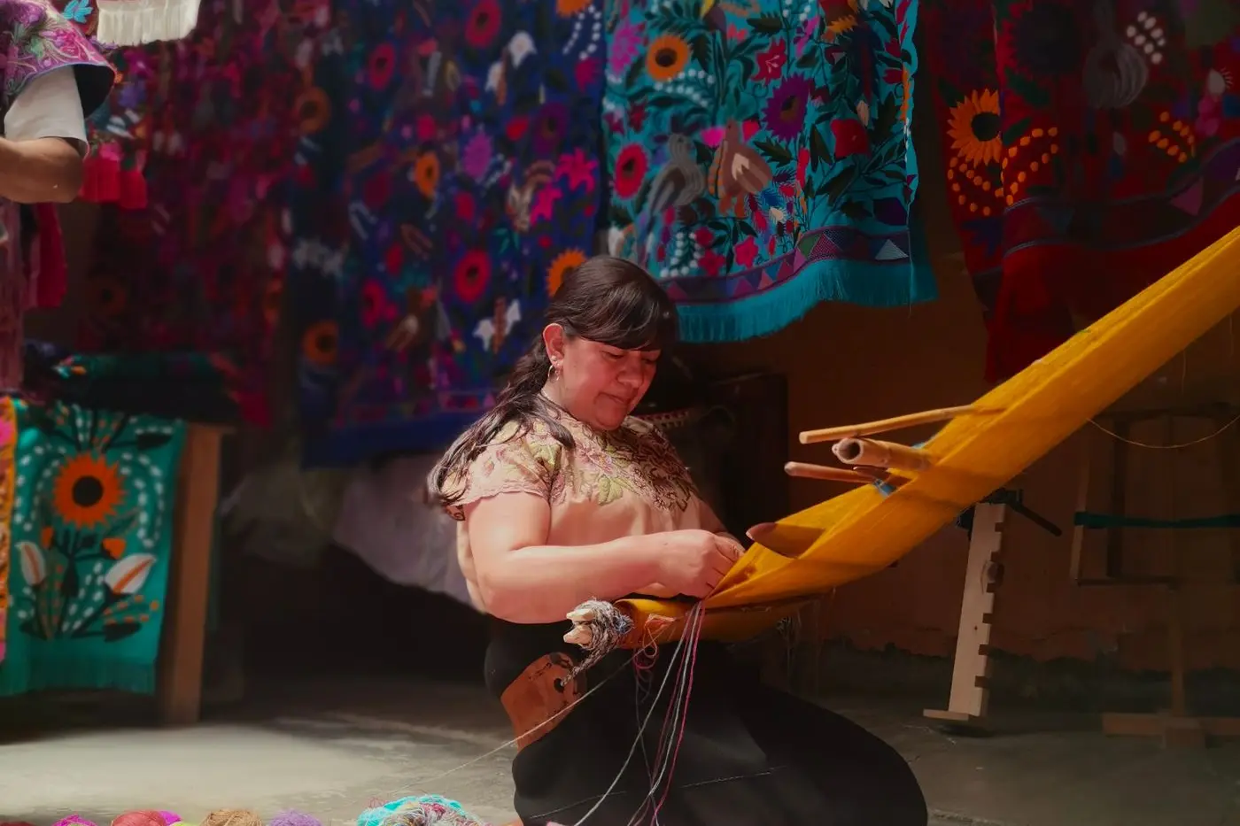 Mujer tzotzil tejiendo en telar de cintura en Zinacantán, con textiles bordados de flores colgados al fondo